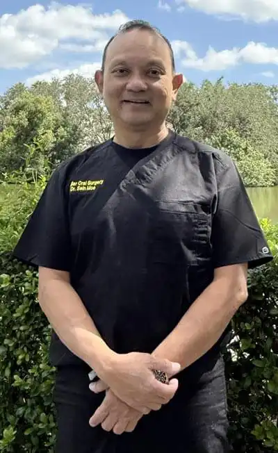Medical professional wearing black surgical scrubs standing outdoors in front of greenery.